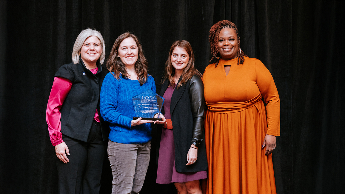 Four women standing together on stage, one holding an award, dressed in various bright outfits.