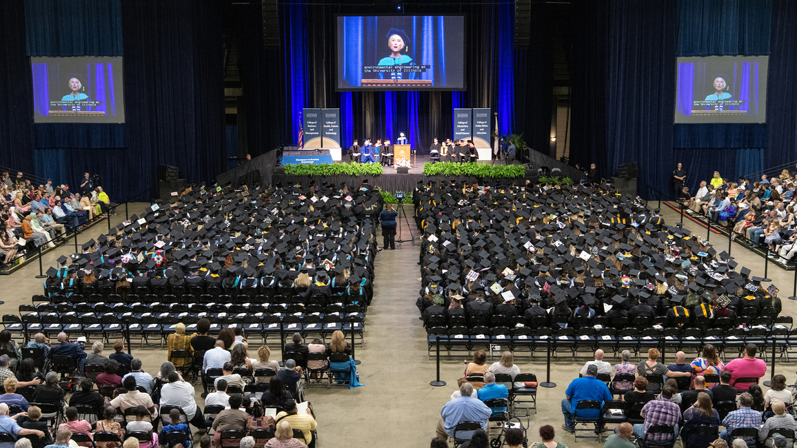 Graduation ceremony with speaker on stage and audience in caps and gowns.