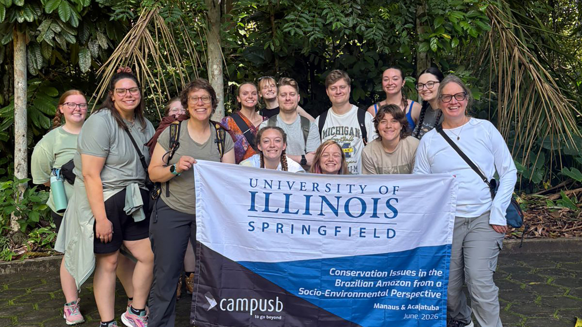 Group of people smiling, holding University of Illinois Springfield banner outdoors.