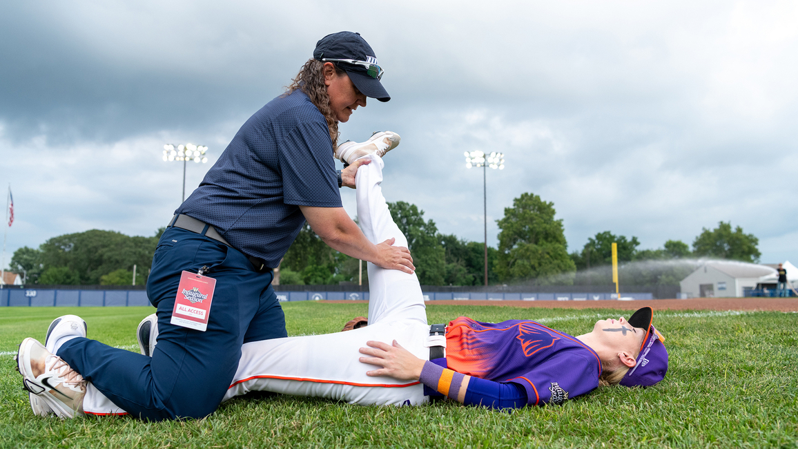 Trainer stretching an athlete's leg on a grassy field under a cloudy sky.