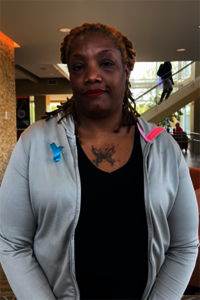 Woman with a neutral expression, wearing a gray hoodie with a blue and pink ribbon, indoors.