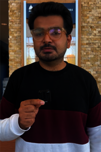 Man in glasses holding a small black object indoors.