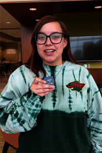 Young person in glasses with sweater, holding a small object indoors, smiling.
