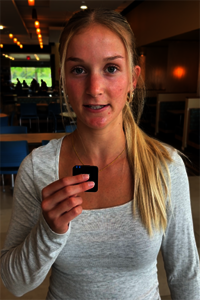 Young woman in a casual setting, holding a small black object, wooden decor in background.