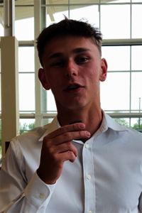 Young man in a white shirt speaking, large windows behind.
