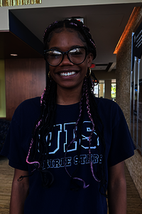 Smiling person with glasses and braided hair, wearing a navy blue shirt indoors.