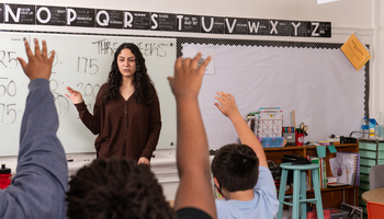 A teacher is shown in a classroom in front of a marker board. Several children's hands are raised to ask a question. 
