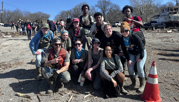 A group of volunteers poses for a photo during an outdoor cleanup event, wearing work gloves, boots, and bandanas. A damaged boat and debris are visible in the background.