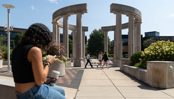 Students walk through the colonnade at the University of Illinois Springfield, while one sits nearby using a phone.