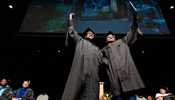 Two graduates in black caps and gowns celebrate on stage by raising their diplomas triumphantly, smiling as they stand side by side. Behind them, faculty members in academic regalia sit in rows and a large screen displays a projection of the moment above them.
