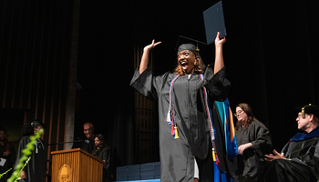 Graduating student celebrates on stage in cap and gown holding diploma at UIS Commencement as faculty applaud.
