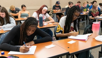 A classroom of students writing in notebooks, laptops open, and water bottles on desks.