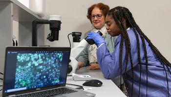A student in a lab coat looks through a microscope while a professor observes beside her, with cell images displayed on a nearby laptop.
