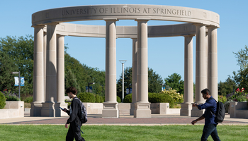 Circular stone structure with tall columns, people walking in front on a sunny day.