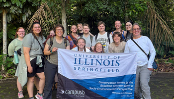Group of people smiling, holding University of Illinois Springfield banner outdoors.