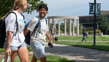 Two students walking on a campus path, smiling, with a stone pavilion in the background.