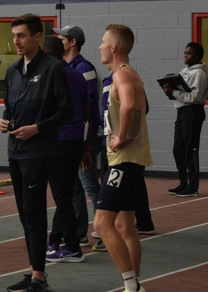 Emmons King in dark blue UIS windbreaker stands with UIS runners inside a track facility.