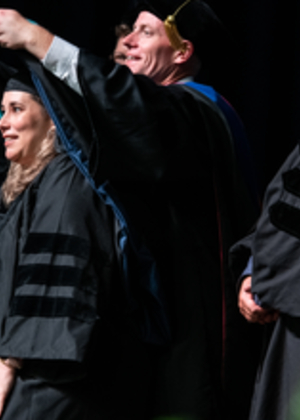 In a ceremonial gesture, the associate dean Travis Bland presents a graduation cap to a beaming graduate, marking the completion of their studies.