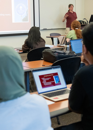 The backs of heads of students sitting in a classroom listing to a professor teach. Several laptop computers are visible.