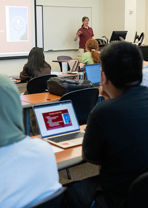 The backs of heads of students sitting in a classroom listing to a professor teach. Several laptop computers are visible.