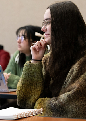 A student in class smiling with a notebook on the desk and pen in hand.