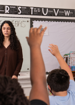 A teacher is shown in a classroom in front of a marker board. Several children's hands are raised to ask a question. 