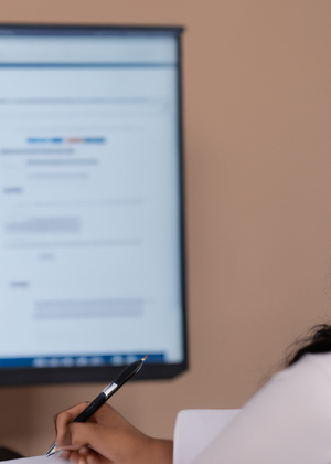 A person from behind sitting at a desk and holding a pen in front of a blurred computer screen.