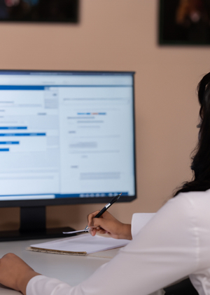 A person from behind sitting at a desk and holding a pen in front of a blurred computer screen.