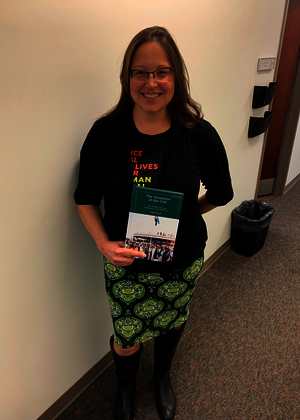 Dr. Kristi Barnwell, a white woman in a black sweater and green dress, smiling and holding a copy of her new book. 