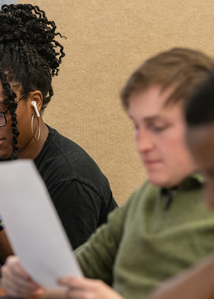 Three students sitting at a desk in a classroom reading pieces of paper. 
