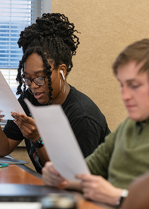 Two students sitting at a desk in a classroom reading pieces of paper. 