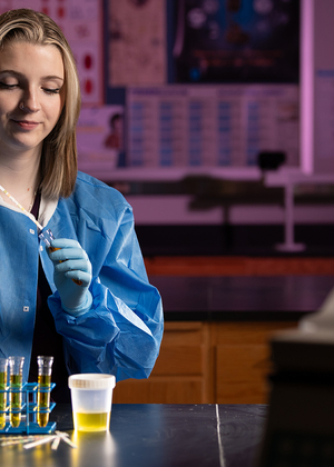 An MLS student wearing a blue lab coat suctions a liquid from a vial.