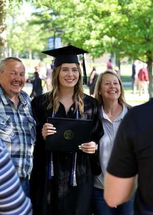 A woman in regalia stands flanked by a man and a woman, posing for a photo with her diploma cover.
