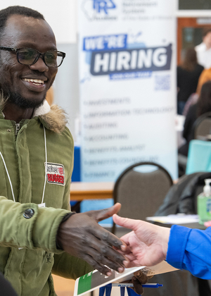 A smiling job fair attendee shakes hands with a recruiter at a table with a "We're Hiring" sign in the background.