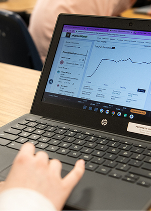 A laptop with stock trading information on the screen sitting on a desk with a person's hand above the mousepad. 