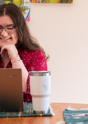 Student smiling while working on a laptop at a table with a tumbler, colorful art hanging on the wall behind her.