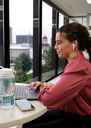 Young woman works on a laptop at a high-rise office window with earbuds, water bottle, and phone nearby.