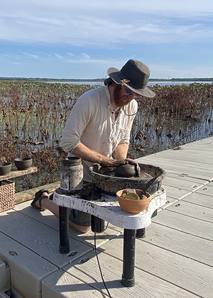 A person shapes clay on a pottery wheel outdoors on a dock at the Emiquon Preserve, surrounded by wetland plants and water.