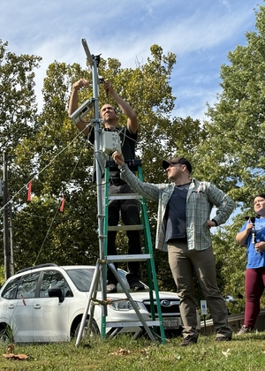 Three researchers assembling weather monitoring station on ladder with atmospheric sensors in parking area