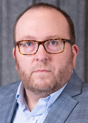 Man with glasses in a blue plaid suit, serious expression, gray background.
