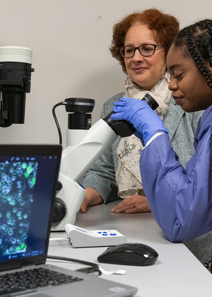 A student in a lab coat looks through a microscope while a professor observes beside her, with cell images displayed on a nearby laptop.