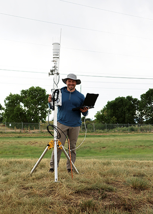 Man with tablet stands by weather station in grassy field.