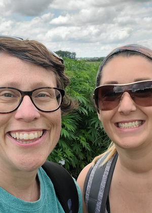 Two smiling people outdoors, cloudy sky, green foliage in the background.