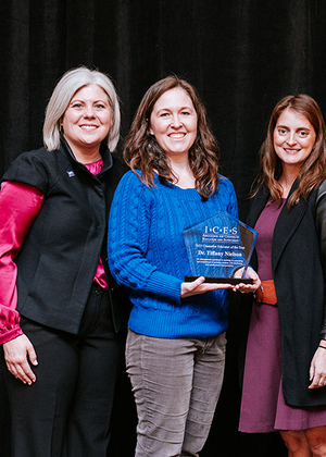 Three women smiling, one holding an award in a blue sweater. Black background.