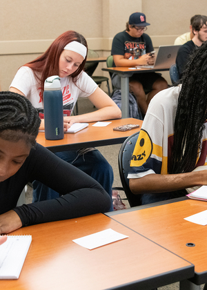 Students attentively writing in a classroom, sitting at desks with notebooks.