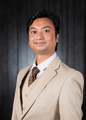 Man in a tan suit with a tie, black background, smiling gently.