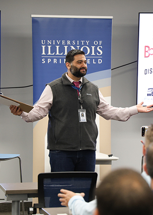 Man speaking at a presentation, University of Illinois Springfield banner behind him.
