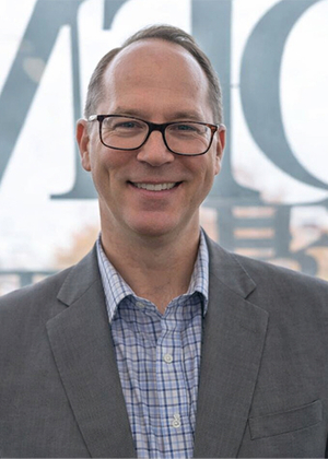 Smiling man in glasses and suit, standing indoors.