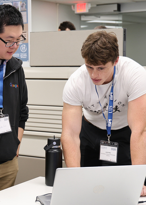 Two men discussing work at a desk with a laptop in an office setting.
