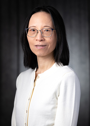 Woman with glasses and a white cardigan, dark background, neutral expression.
